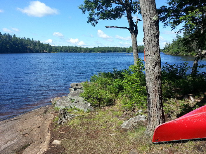 A red canoe sticks into the edge of the frame in a photo taken from the shore looking out over a lake on a sunny day