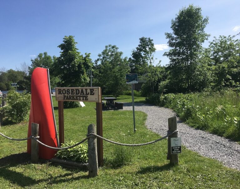 A sign that reads Rosedale Parkette with a half-canoe standing upright beside it and trail leading into the parkette