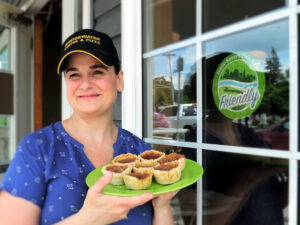 A smiling woman holds up a tray with a half dozen butter tarts on it