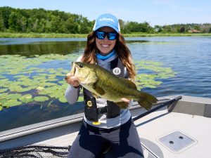 A smiling woman holds a bass towards the camera, lily pads in the background