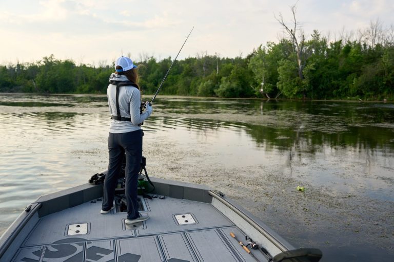 A person fishes from the bow of a boat, with a green shore seen in the background