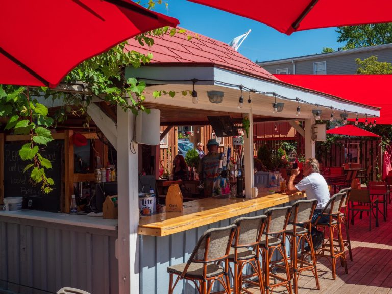 A man in a white t-shirt sits an an outdoor bar, surrounded by red umbrellas