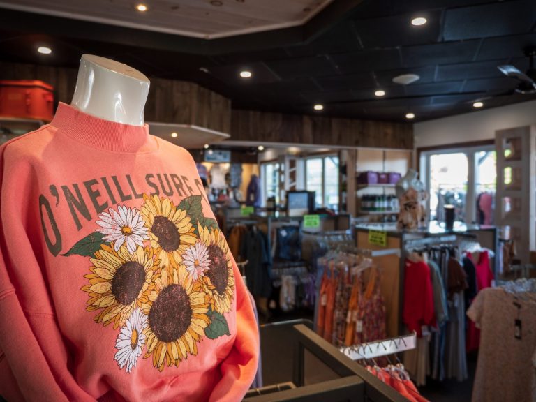 A orange sweatshirt decorated with sunflowers and the words "O'Neill Surf" in the foreground, more racks of apparel in the background