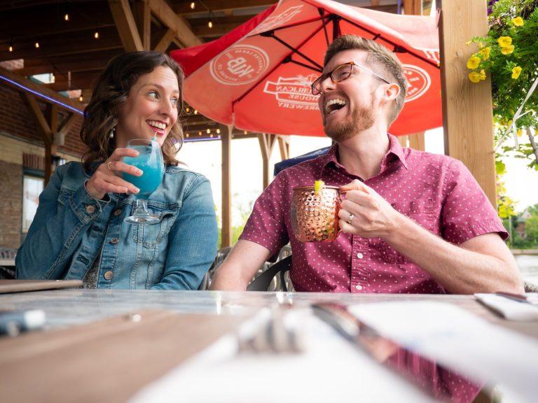 Two people on a patio holding cocktails look at each other while smiling and laughing