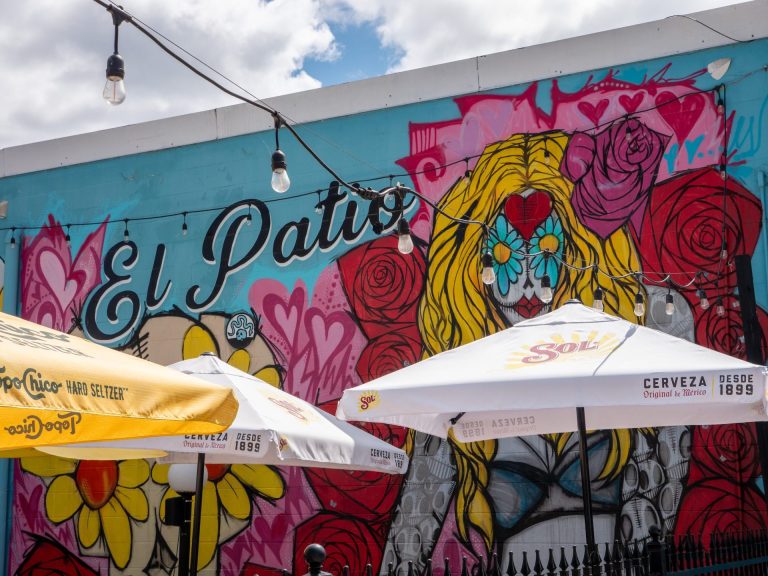 Mexican-influenced mural of a wild-haired character surrounded by flowers, with the words "El Patio" beside it and several patio tables with umbrellas in front of it