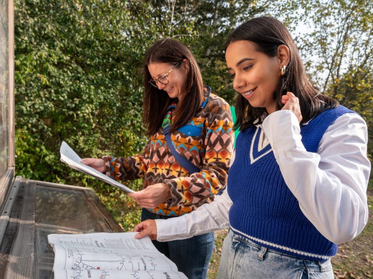 Two people in sweaters stand in front of an outdoor plaque and check a trail map