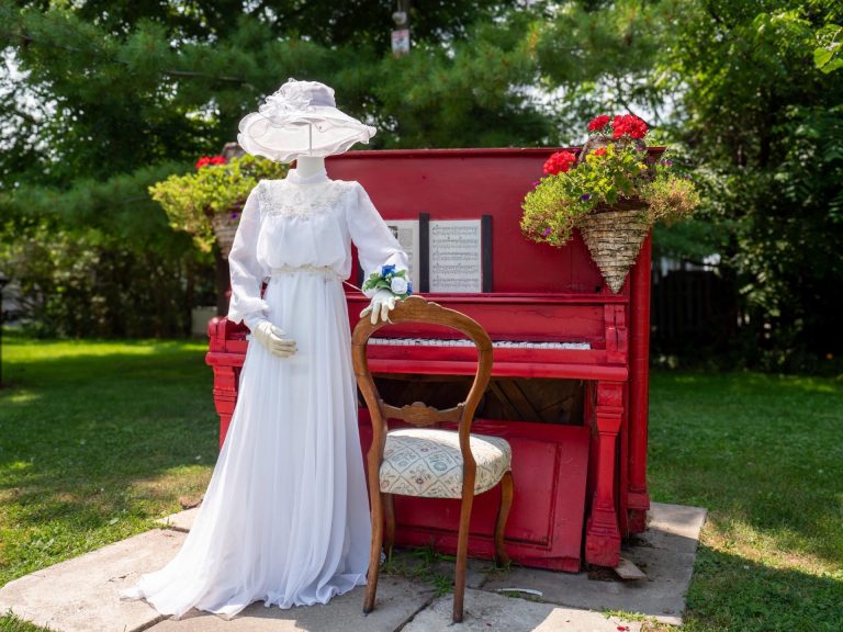 A mannikin in a formal white dress stands beside an outdoor piano that has been painted red