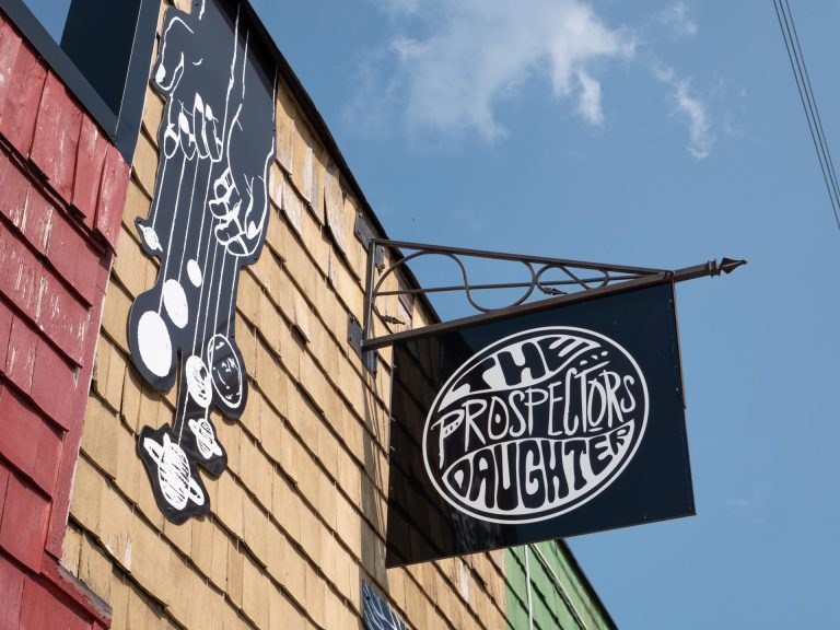A sign reading the "Prospector's Daughter" is framed against a blue sky and cedar shake siding