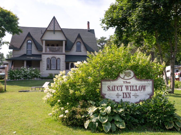 A sign in a bush reads "Saucy Willow Inn," with an historic building with three gables behind it